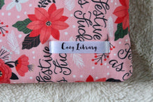 Load image into Gallery viewer, A close up of the Cozy Library tag is shown on the bottom right corner of a Festive AF book sleeve.