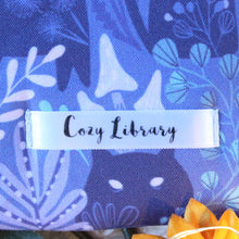 Load image into Gallery viewer, A close up of the Cozy Library tag is show on the bottom right corner of a Purple Forest Guardian book sleeve.