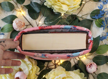 Load image into Gallery viewer, A hand is holding up a medium-sized book sleeve, which contains a thick hardcover book. There are leaves and flowers in the background of the photo.