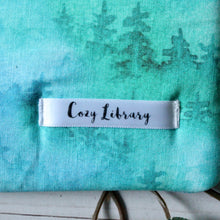 Load image into Gallery viewer, A close up of the Cozy Library tag is shown on the bottom right corner of a Forest book sleeve.