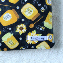 Load image into Gallery viewer, A close up of the Cozy Library tag is shown on the bottom right corner of a Honey Jars book sleeve.