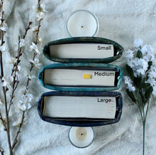 Load image into Gallery viewer, Pictured is the difference in Cozy Library's book sleeves from Small, Medium, to Large, from above. Width of the book's are visible. The sleeves are being held up by 2 candles so they won't fall over.