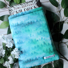 Load image into Gallery viewer, A hardcover book of "Children of Blood and Bone" by Tomi Adeyemi is shown halfway inside a medium-sized Forest book sleeve. The designed fabric shows a green-blue foggy forest in a watercolour-paint style. Also shown beneath the product are leaves on a white-washed wood background. One bunch of white flowers rest beside the book sleeve. Photo is titled to the left.