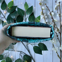 Load image into Gallery viewer, Pictured from a bird's-eye-view, is a hand holding up a medium-sized Green Forest Guardian book sleeve. Inside the book sleeve is the book titled "Children of Blood and Bone" by Tomi Adeyemi. In the background of the photo are leaves and white flowers, accompanied by fairy lights.