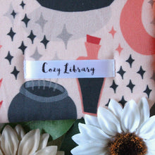 Load image into Gallery viewer, A close up of the Cozy Library tag is shown on the bottom right corner of a Cozy Occult book sleeve.