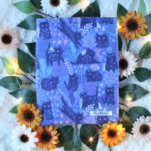 Load image into Gallery viewer, Pictured is a medium-sized Purple Forest Guardian book sleeve. In the designed fabric are cats which are seen looking towards the viewer. There are white, lilac, light green, and purple woodland/forest flora. There are tiny long-haired folk either beside, or on top of some cats. In the background of the photo are white, beige and orange flowers, and green leaves, as well as a stand of fairy lights--all on top of a white textured background.