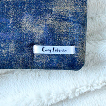 Load image into Gallery viewer, A close up of the Cozy Library tag is shown on the bottom right corner of a Indigo Gold Dust book sleeve.