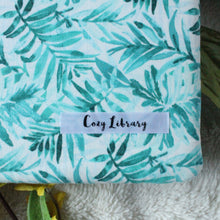 Load image into Gallery viewer, A close up of the Cozy Library tag is shown on the bottom right corner of a Blue Tropical Palm book sleeve.