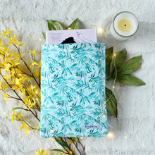 Load image into Gallery viewer, A small Blue Tropical Palm book sleeve is pictured. In the book sleeve is a paperback book titled "Emergency Contact" by H.K. Choi. Blue watercolured-style painted Palm leaves is the design on the fabric, in a repeat pattern style. Also pictured is one bunch of yellow flowers, green leaves, a small candle, for size comparison. Fairy lights in the photo give a fantasy-esque feel to the picture.