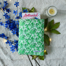 Load image into Gallery viewer, A small Green Tropical Palm book sleeve is carrying a book called "Antisocial" by Heidi Cullinan. There are blue flowers, leaves and an unlit candle in the background.