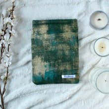 Load image into Gallery viewer, Green Gold Dust has splashes of green, teal, and gold in an abstract vision. Green Gold Dust book sleeve sits spaced between a bunch of white flowers to the left, and 3 varying sizes of candles to the right.