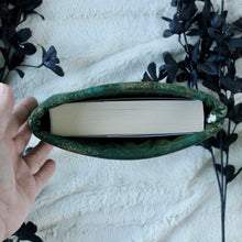Load image into Gallery viewer, A hand is holding up a small-sized Green Gold Dust book sleeve, which holds a Emergency Contact, by Mary H.K. Choi. There are black flowers in the background.