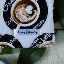 Load image into Gallery viewer, A close up of the Cozy Library tag is shown on the bottom right corner of a Coffee Shop book sleeve.