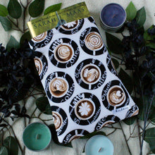 Load image into Gallery viewer, A large coffee-themed book sleeve is pictured. In the fabric design shows coffee art, with different kinds of coffee as words in white text, on a black circled background. Within the book sleeve is a hardcover copy of "Kingdom of Ash" by Sarah J. Maas. There are candles and black flowers in the background of the photo.