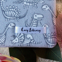 Load image into Gallery viewer, A close up of the Cozy Library tag is shown on the bottom right corner of a Dinos book sleeve.