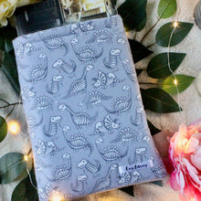 Load image into Gallery viewer, A hardcover copy of "An Ember in the Ashes" by Sabaa Tahir is inside a medium sized Dino book sleeve. There are leaves and fairy lights in the background of the photo.