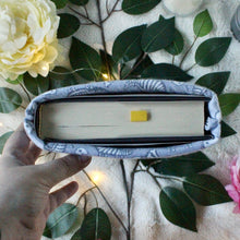 Load image into Gallery viewer, A hand is shown holding up a medium-sized Dinos book sleeve, which has "An Ember in the Ashes" inside it. There are leaves in the background.