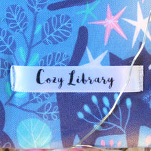 Load image into Gallery viewer, A close up of the Cozy Library tag is shown on the bottom right corner of a Blue Forest Guardian book sleeve.
