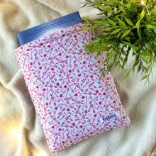 Load image into Gallery viewer, A Valenitine&#39;s Day themed fabric, which has red and pink arrows through hearts are scattered in a repeated fashion. The background colour of the fabric is white. A hardcover copy of &quot;Circle of Shadows&quot; is peeking out of the sleeve. There&#39;s a plant for size comparison.
