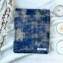 Load image into Gallery viewer, There is a blue, indigo, and gold dust fabric that makes Indigo Gold Dust book sleeve. There are white flowers on the left side of the photo. On the right side, there are three white candles in 3 various sizes on the right side of the picture. 
