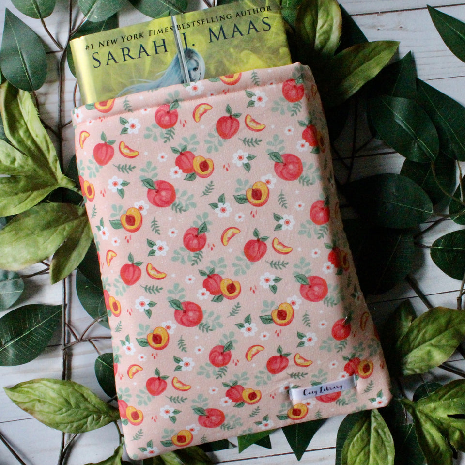 A hardcover book sits within a large-sized Just Peachy book sleeve.There are green leaves in the background.