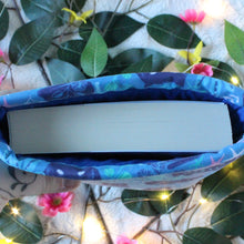 Load image into Gallery viewer, A book is being shown inside a Small sized Blue Forest Guardian book sleeve. Pictured from bird&#39;s eye view, so you can see how to expect your book to fit.In the background there are pink and yellow flowers, accompanied by green leaves, and fairy lights.
