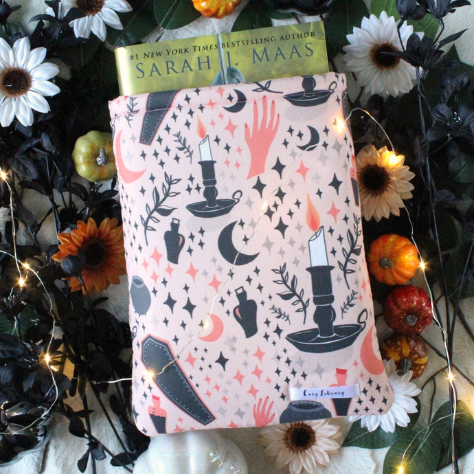 In the fabric design of the Cozy Occult book sleeve are candles in candlesticks, a peach hand with red nail-polish, potion bottles, coffins, and grey and black diamon-stars. A light peach background in the fabric brings everything together. A large-sized Cozy Occult book sleeve is shown. Within the book sleeve is a hardcover copy of 