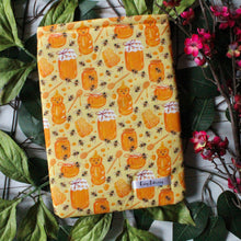 Load image into Gallery viewer, A small-sized honey-themed book sleeve. The fabric is designed as various jars of honey, a teddy-bear squeeze bottle filled with honey, a honey spoon with honey on it, a honey comb, splatters of honey, and bees are shown in a repeat pattern.
