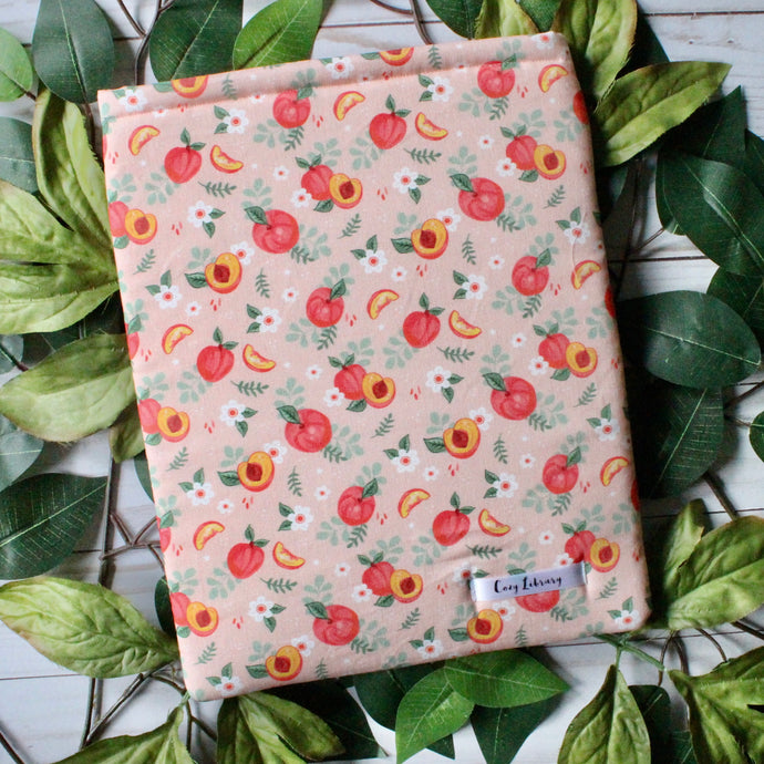 painted peaches, whole, cut in half, and sliced peaches decorate this fabric design. There's a soft peach background colour in the fabric. Beneath the book sleeve are two-toned green leaves.