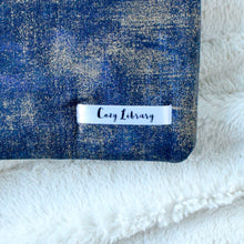 Load image into Gallery viewer, A close up of the Cozy Library tag is shown on the bottom right corner of a Indigo Gold Dust book sleeve.
