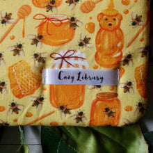 Load image into Gallery viewer, A close up of the Cozy Library tag is shown on the bottom right corner of a Dreaming of Honey book sleeve.

