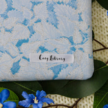 Load image into Gallery viewer, A close up of the Cozy Library tag is shown on the bottom right corner of a Periwinkle book sleeve.
