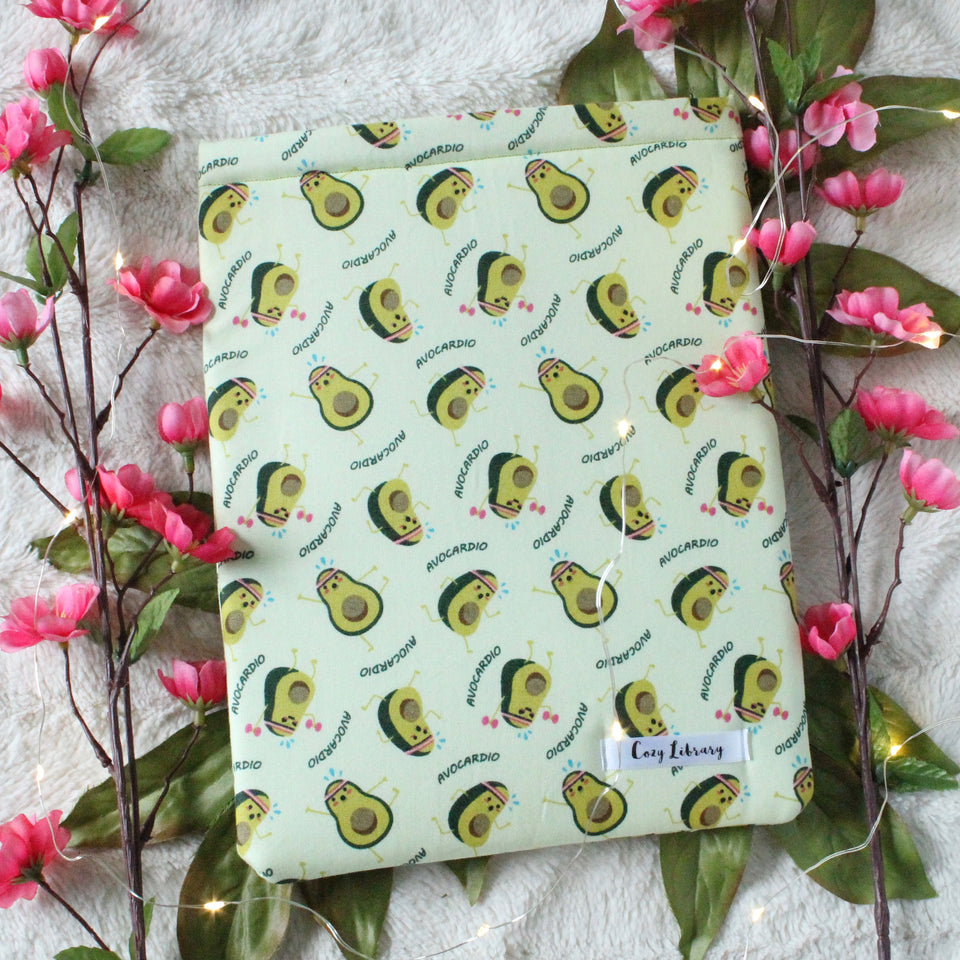 A medium-sized book sleeve is pictured. The design of the book sleeve is half an Avocado, seed in, with arms and legs, doing cardio. The word "Avocardio" is placed throughout this repeat design. Background fabric colour is a soft light green. Also pictured are two pink flower bunches, one on either side of the book sleeve. As well as fairy lights, which add warmth to the photo.