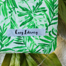 Load image into Gallery viewer, A close up of the Cozy Library tag is shown on the bottom right corner of a Green Tropical Palm book sleeve.
