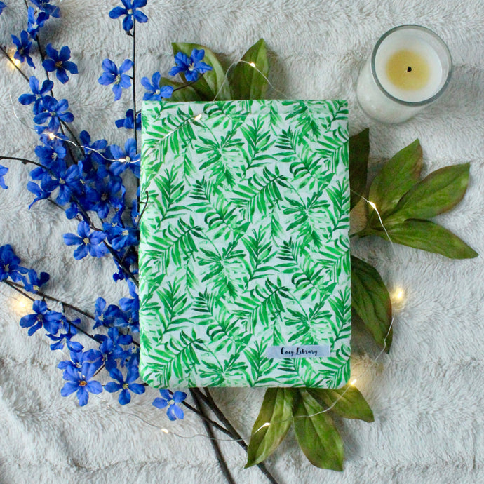 Green watercoloured palm leaves in a repeat pattern make up the Green Tropical Palm book sleeve. In the background there are leaves, blue flowers, an unlit candle, and fairy lights.