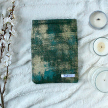 Load image into Gallery viewer, Green Gold Dust has splashes of green, teal, and gold in an abstract vision. Green Gold Dust book sleeve sits spaced between a bunch of white flowers to the left, and 3 varying sizes of candles to the right. 
