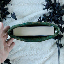 Load image into Gallery viewer, A hand is holding up a small-sized Green Gold Dust book sleeve, which holds a Emergency Contact, by Mary H.K. Choi. There are black flowers in the background.
