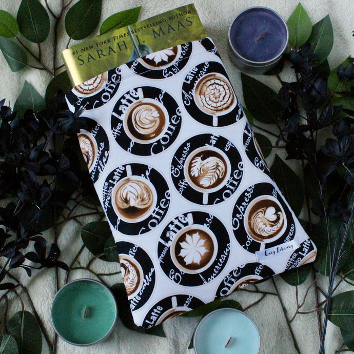 A large coffee-themed book sleeve is pictured. In the fabric design shows coffee art, with different kinds of coffee as words in white text, on a black circled background. Within the book sleeve is a hardcover copy of 