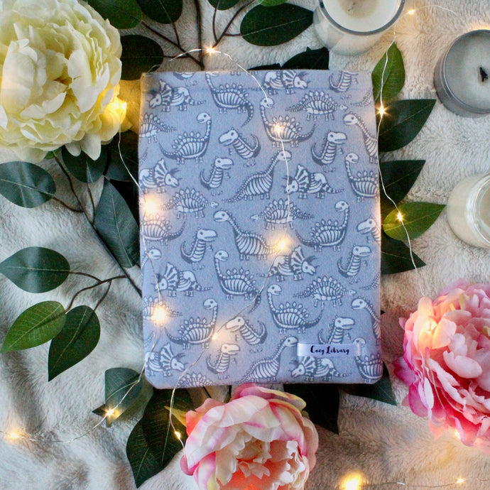 A medium-sized book sleeve, wherein the fabric shows cartoon style dinosaurs' bones in white, with a dark grey silhouette body to match. The background colour is a light grey to tie everything together.There are fairylights atop the Dino book sleeve. Leaves, flowers and unlit candles make the background of the photo. 