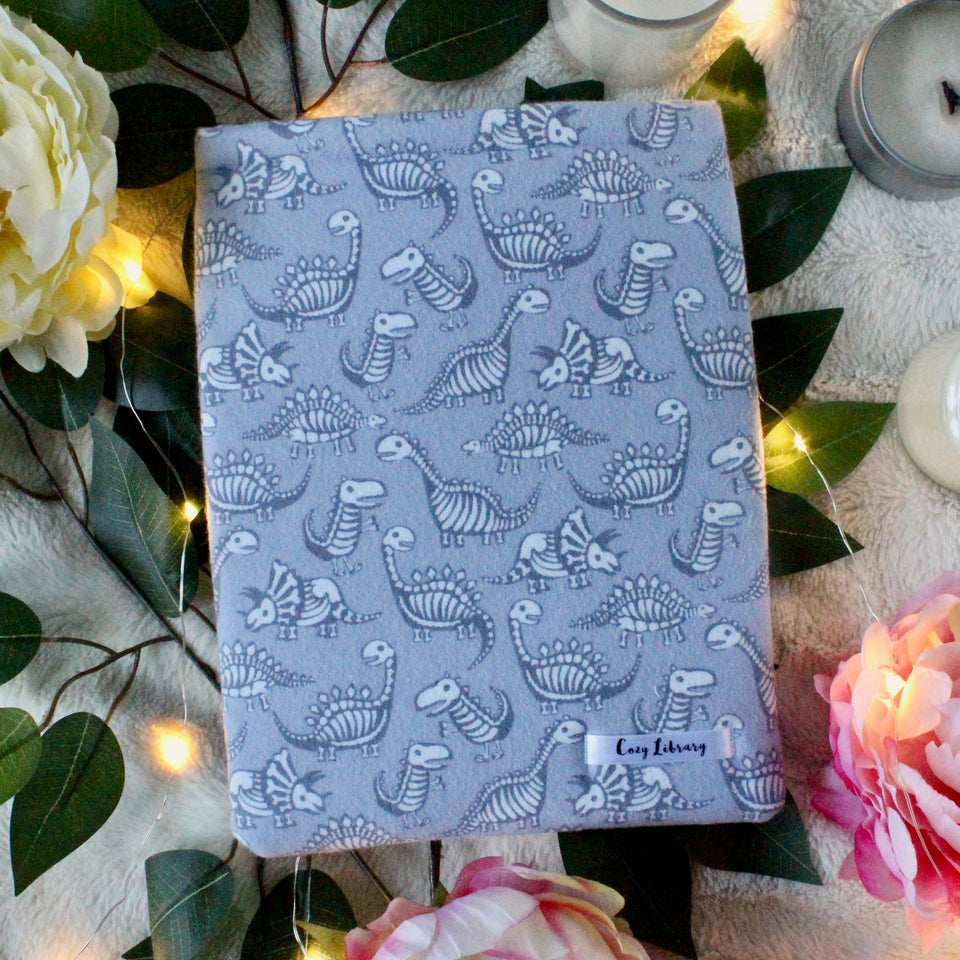 A medium-sized book sleeve, wherein the fabric shows cartoon style dinosaurs' bones in white, with a dark grey silhouette body to match. The background colour is a light grey to tie everything together. There are fairy lights beneath the Dino book sleeve. Leaves, flowers and unlit candles make the background of the photo. 