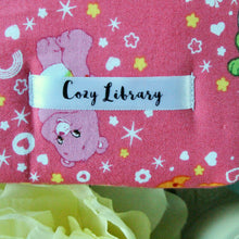 Load image into Gallery viewer, A close up of the Cozy Library tag is shown on the bottom right corner of a Care Bear book sleeve.
