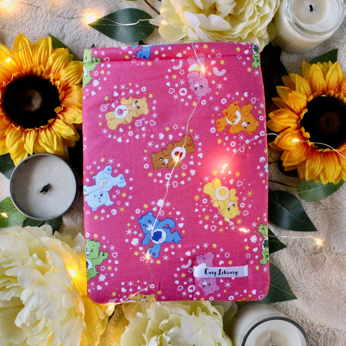 Pictured is a small-zised Care Bear book sleeve. Designed are various deifferent Care Bears in different positions being happy. The background colour of the design is bright pink. Also pictured are candles, yellow and orange flowers, and leaves. Fairy Lights are placed throughout the photo which emits a warm light.
