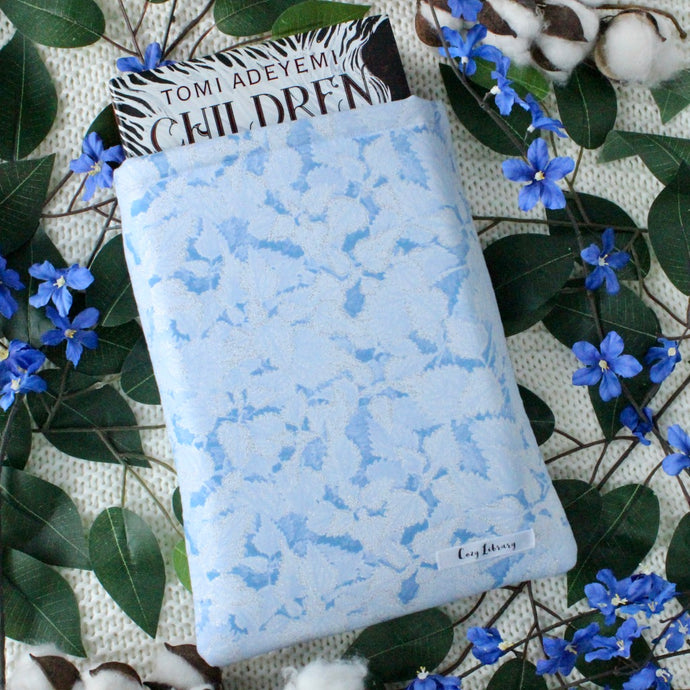 Periwinkle is captured, with a book halfway into a book sleeve. There are blue flowers, green leaves, and cotton in the pictures. The background is a textured white blanket.