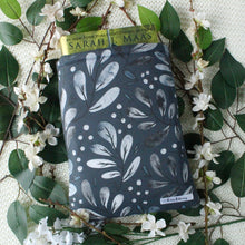 Load image into Gallery viewer, A thick book is shown partially inside a Snowberries book sleeve. In the photo is also pictured white flowers, and green leaves.
