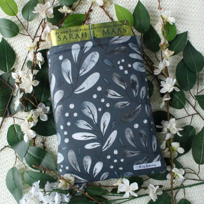A thick book is shown partially inside a Snowberries book sleeve. In the photo is also pictured white flowers, and green leaves.