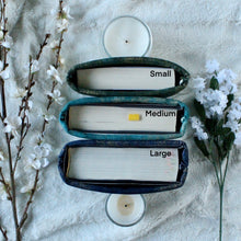 Load image into Gallery viewer, Pictured is the difference in Cozy Library&#39;s book sleeves from Small, Medium, to Large, from above. Width of the book&#39;s are visible. The sleeves are being held up by 2 candles so they won&#39;t fall over.
