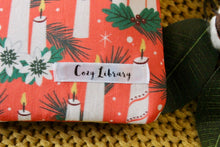 Load image into Gallery viewer, A close up of the Cozy Library tag is shown on the bottom right corner of a Candles Aglow book sleeve.
